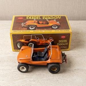 May include: An orange Cox Dune Buggy toy vehicle with black seats and a steering wheel, positioned in front of its original yellow and brown cardboard box. The box features illustrations of the buggy and the text Dune Buggy, Cox, and Throttle Equipped.