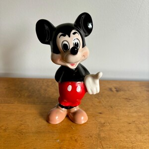 Puede incluir: Una figurita de cer&aacute;mica negra y blanca de Mickey Mouse. Mickey lleva sus pantalones cortos rojos caracter&iacute;sticos con dos botones blancos y guantes blancos. Est&aacute; sonriendo y tiene una nariz negra y orejas grandes negras.