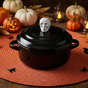 May include: A round, black cooking pot with two side handles and a matching lid. The lid features a white, three-dimensional skull-shaped handle centered on top. The pot has a smooth, glossy finish and is constructed with a circular base and sides.