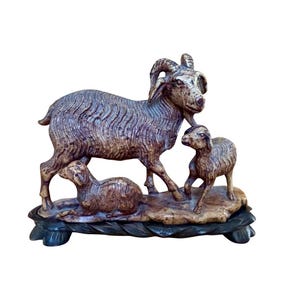 May include: A detailed sculpture of a ram with two lambs. The ram stands tall with curved horns, while the lambs are positioned at its feet. The sculpture is in shades of brown and beige, mounted on a dark base.