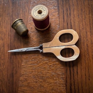 May include: A pair of small sewing scissors with light-colored handles, a brass thimble with a textured surface, and a wooden spool of dark maroon thread labeled Finest Silk Est. 1890. These sewing tools are arranged together on a flat surface.