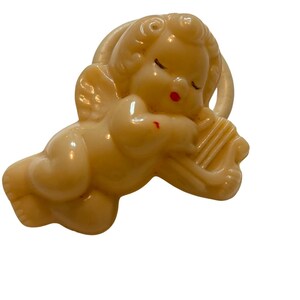 May include: A cream-colored plastic brooch shaped like a sleeping cherub with curly hair and wings. The cherub is resting its head on a small harp. The figure features subtle red painted details on the lips and a small mark on the shoulder.