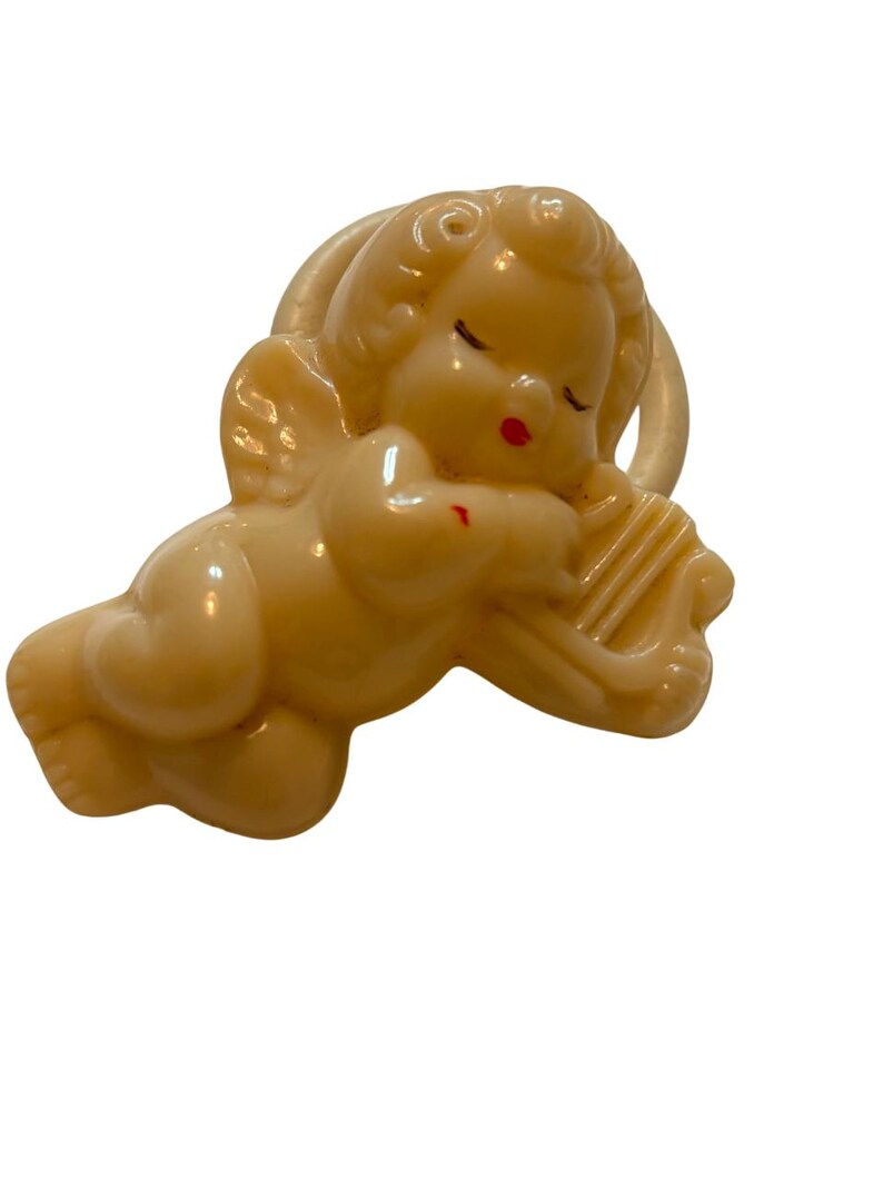 May include: A cream-colored plastic brooch shaped like a sleeping cherub with curly hair and wings. The cherub is resting its head on a small harp. The figure features subtle red painted details on the lips and a small mark on the shoulder.