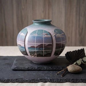 May include: A round ceramic vase with a light purple body and a teal rim. The vase features a landscape design with a window grid pattern. The landscape depicts a coastal scene with mountains, water, and sky. The vase sits on a dark gray slate.