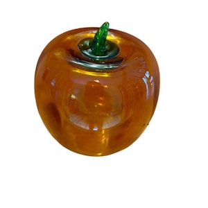 May include: A glass apple sculpture with a shiny, amber-colored body and a green stem. The apple has a smooth, rounded shape and reflects light, creating a glossy appearance. The stem is a vibrant green, contrasting with the amber body.