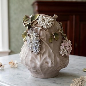 May include: A beige ceramic vase with a textured surface and floral embellishments. The vase features clusters of white, pink, and blue flowers with green leaves. The vase is sitting on a marble surface.