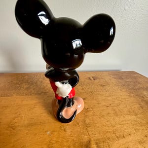 Puede incluir: Una figurita de cer&aacute;mica negra y roja de Mickey Mouse. Mickey est&aacute; de pie con la mano derecha en la cadera y la mano izquierda levantada. Lleva sus caracter&iacute;sticos pantalones cortos rojos y zapatos amarillos.
