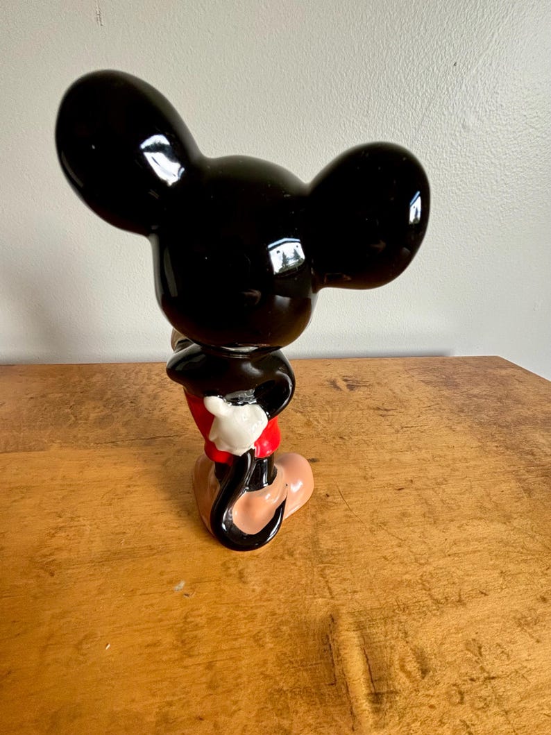 Puede incluir: Una figurita de cer&aacute;mica negra y roja de Mickey Mouse. Mickey est&aacute; de pie con la mano derecha en la cadera y la mano izquierda levantada. Lleva sus caracter&iacute;sticos pantalones cortos rojos y zapatos amarillos.