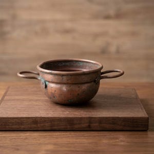 May include: A small, antique copper bowl with two handles. The bowl has a rounded shape and a weathered, aged appearance, with visible patina and discoloration. The bowl is sitting on a wooden surface.