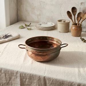 May include: A copper bowl with two handles sits on a white tablecloth. The bowl has a weathered appearance with a dark interior. Other kitchen items are in the background, including wooden spoons, small plates, and a ceramic jar.