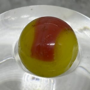 May include: A colorful glass marble with a yellow base and a red rectangular design. The marble is set in a clear, shallow dish. The marble's surface is smooth and reflective, with a slight sheen.