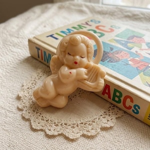 May include: A cream-colored plastic cherub figurine playing a harp, featuring molded curly hair, closed eyes, and red painted lips. The cherub is positioned in front of a vintage children's book titled Timmy's ABCs with colorful illustrations.