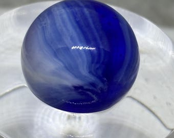 May include: A blue and white swirled glass marble. The marble is a deep blue color with white streaks and swirls throughout. The marble is sitting on a clear, glass surface.