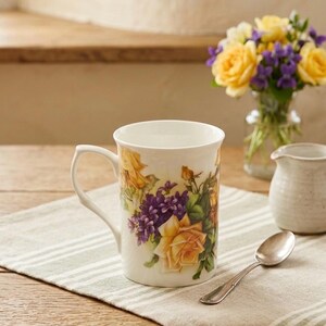 May include: A white ceramic mug featuring a floral pattern of yellow roses and purple flowers. The mug has a curved handle and a smooth finish. It is positioned on a light-colored fabric surface next to a small silver spoon and a ceramic pitcher.