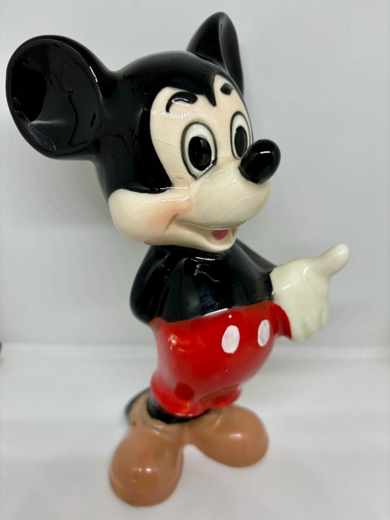 Puede incluir: Una figurita de cer&aacute;mica de Mickey Mouse. La figurita es negra y blanca con pantalones cortos rojos y botones blancos. Mickey est&aacute; sonriendo y tiene la mano derecha levantada.