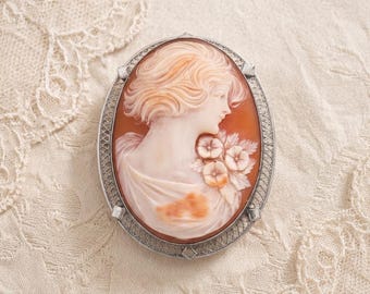 May include: An oval brooch featuring a carved shell cameo of a person in profile with wavy hair and flowers on their shoulder. The cameo is set in a silver-toned metal frame with a delicate, openwork lattice pattern around the outer edge.