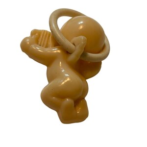 May include: A light peach-colored plastic baby pacifier featuring a molded cherub figure attached to a circular ring handle. The cherub is depicted in a kneeling position, with one arm raised toward the ring, showcasing a smooth, glossy finish.