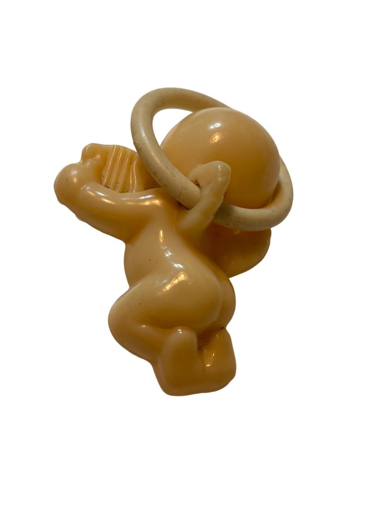 May include: A light peach-colored plastic baby pacifier featuring a molded cherub figure attached to a circular ring handle. The cherub is depicted in a kneeling position, with one arm raised toward the ring, showcasing a smooth, glossy finish.
