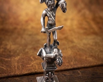 May include: A silver-toned metal figurine depicting a standing figure wearing a winged helmet, holding a sword in one hand and a severed head in the other, positioned atop a rectangular pedestal base featuring a fleur-de-lis emblem detail.