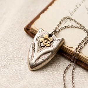 May include: A silver-toned shield-shaped pendant featuring an embossed floral pattern and a small gold-toned flower accent at the top. The pendant hangs from a delicate silver-toned chain, showcasing intricate etched details across its surface.