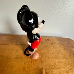 Puede incluir: Una figurita de cer&aacute;mica de Mickey Mouse de pie con la mano derecha en la cadera. Lleva pantalones cortos rojos, zapatos negros y tiene orejas negras y una nariz negra.