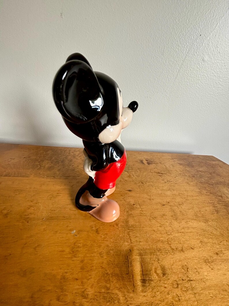Puede incluir: Una figurita de cer&aacute;mica de Mickey Mouse de pie con la mano derecha en la cadera. Lleva pantalones cortos rojos, zapatos negros y tiene orejas negras y una nariz negra.