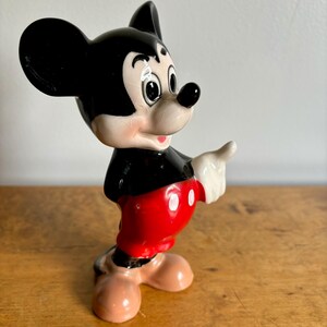 Puede incluir: Una figurita de cer&aacute;mica de Mickey Mouse. Est&aacute; de pie con el brazo derecho extendido y se&ntilde;alando con el dedo. Lleva sus caracter&iacute;sticos pantalones cortos rojos con botones blancos y zapatos negros.
