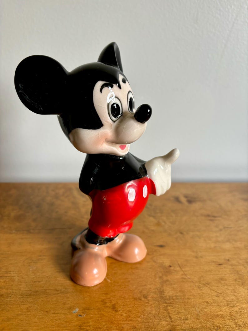 Puede incluir: Una figurita de cer&aacute;mica de Mickey Mouse. Est&aacute; de pie con el brazo derecho extendido y se&ntilde;alando con el dedo. Lleva sus caracter&iacute;sticos pantalones cortos rojos con botones blancos y zapatos negros.