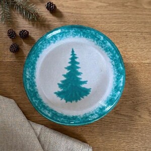 May include: A round ceramic plate featuring a white center with a teal evergreen tree illustration and a wide, textured teal border. The plate has a smooth, glossy finish and a simple, circular design that highlights the central tree motif.