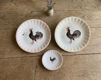 May include: Two large and one small round ceramic plates with fluted edges, each featuring a central illustration of a black rooster with red accents. The plates have a cream-colored base with visible signs of wear and distressed paint finish.