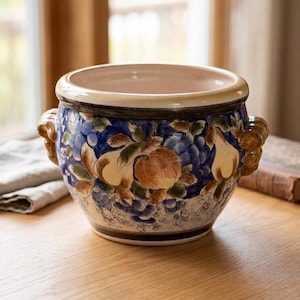 May include: A ceramic planter with a wide, cream-colored rim and two textured, brown side handles. The exterior features a hand-painted design of blue grapes, yellow pears, and brown apples against a deep blue background with green leaves.