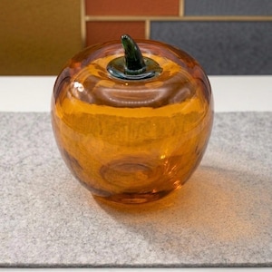 May include: A translucent amber-colored glass apple sculpture with a smooth, rounded body and a dark green glass stem attached to the top. The object features a glossy finish that reflects light, creating highlights across its curved surface.