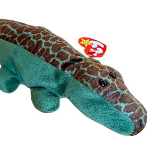 May include: A plush alligator toy with a green body and a brown, patterned back. The toy has black eyes and a red tag attached. The alligator is lying down on a white background.