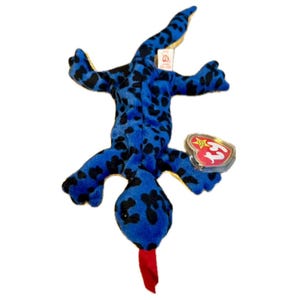 May include: A blue plush lizard toy with black spots and a red tongue. The toy has a tag attached with a heart-shaped emblem. The lizard is lying on its back, with its legs and tail extended.
