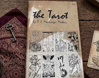 May include: A beige paperback book titled the Tarot by S. L. MacGregor Mathers with a grid of eight black and white tarot card illustrations on the cover. The book is placed on a dark red patterned cloth next to a small metal skeleton key.