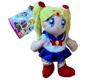 May include: A plush doll of a character with blonde hair styled in pigtails, red accents, and blue eyes. The doll wears a sailor-style outfit with a white top, blue skirt, and red boots. A tag with illustrations is attached.