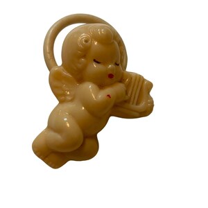 May include: A cream-colored plastic cherub figurine featuring a halo behind its head and wings on its back. The cherub has closed eyes, red painted lips, and a small red mark on its arm while holding a small harp instrument in its hands.