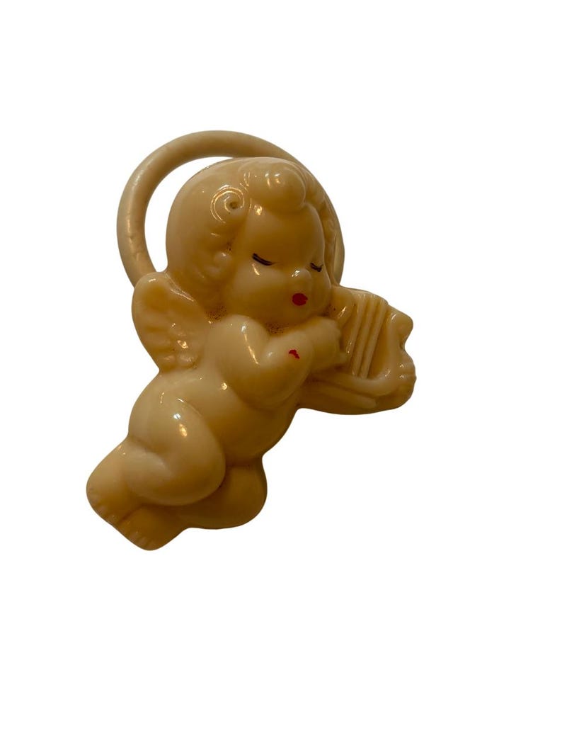 May include: A cream-colored plastic cherub figurine featuring a halo behind its head and wings on its back. The cherub has closed eyes, red painted lips, and a small red mark on its arm while holding a small harp instrument in its hands.