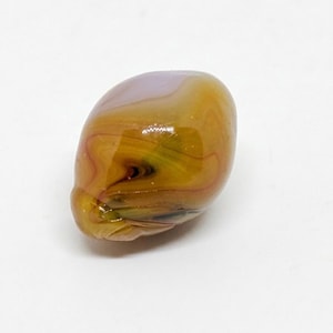 May include: A smooth, rounded glass bead featuring a swirling pattern of amber, gold, and brown tones with a subtle hint of lavender at the top. The surface is polished and reflective, showcasing organic, flowing lines throughout the material.
