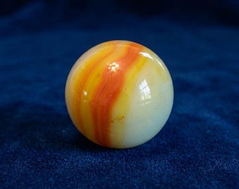 May include: A single, round marble with a swirl of orange, yellow, and white colors. The marble has a glossy finish and reflects light. The marble is a classic toy.