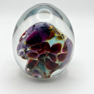 May include: A clear glass paperweight with an internal design of swirling colors. The colors include deep purple, burgundy, teal, and gold. The paperweight is egg-shaped and sits on a white surface.
