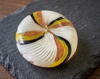 May include: A round glass paperweight featuring a swirling pattern of opaque white, translucent yellow, and metallic copper-toned glass ribbons, accented by thin black lines that radiate from the center toward the outer edge of the object.