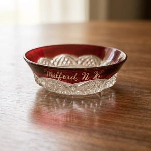 May include: A small, round glass bowl featuring a deep red rim and a clear, faceted base. The red section is engraved with the white script text Milford, N.H. The bowl has a decorative, pressed pattern around its exterior, combining smooth colored glass with intricate, textured clear glass elements.
