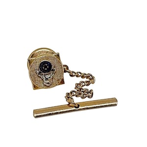 May include: Gold-toned tie tack featuring a square design with a textured surface and a bull's head emblem. The tack includes a chain and a bar for securing to a tie. The bull's head has a dark blue stone in the center.