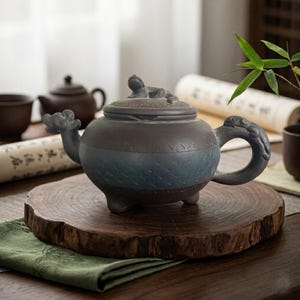 May include: A handcrafted ceramic teapot with a dark brown and blue color scheme. The teapot features a dragon-shaped handle and spout, and a small figure on the lid. It sits on a wooden slab, suggesting a focus on tea ceremony.