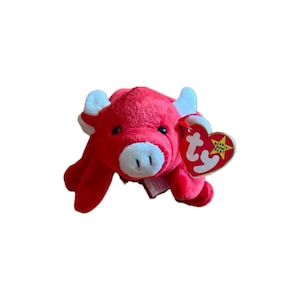 May include: A red plush toy bull with white horns and a white snout. The toy has a Ty Beanie Baby tag attached. The bull is lying down on a white background.
