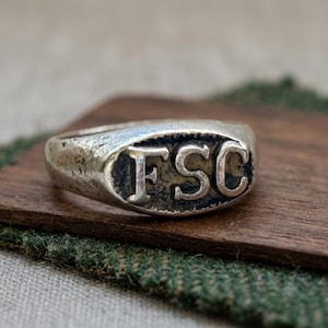 May include: A silver-toned metal ring featuring a textured, weathered surface and an oval face. The center of the ring displays the embossed capital letters F, S, and C in a serif font, set against a darkened, recessed background.