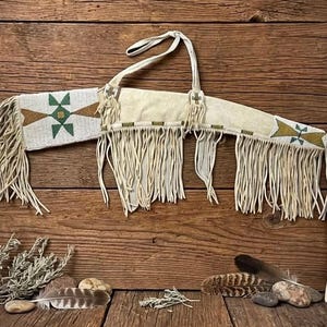 Può includere: Una faretra in pelle color crema con lunghe frange e intricate perline in motivi geometrici bianchi, verdi e dorati. La faretra include una tracolla in pelle ed è decorata con piccole croci di perline in corrispondenza degli attacchi.