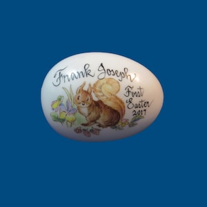 May include: White porcelain egg-shaped ornament with a hand-painted squirrel and flowers. The text "Frank Joseph First Easter 2017" is written on the ornament.