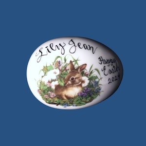 May include: White ceramic Easter egg with a brown bunny rabbit surrounded by flowers and green grass. The egg is decorated with the text "Lily Jean" and "Happy Easter 2024".